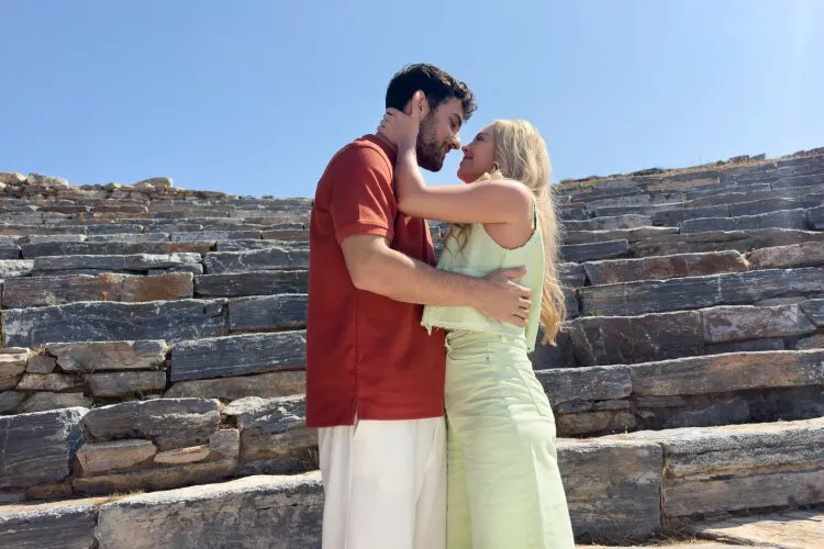 Abby and Theo sharing a romantic moment, embracing in an ancient amphitheater under the blue sky.