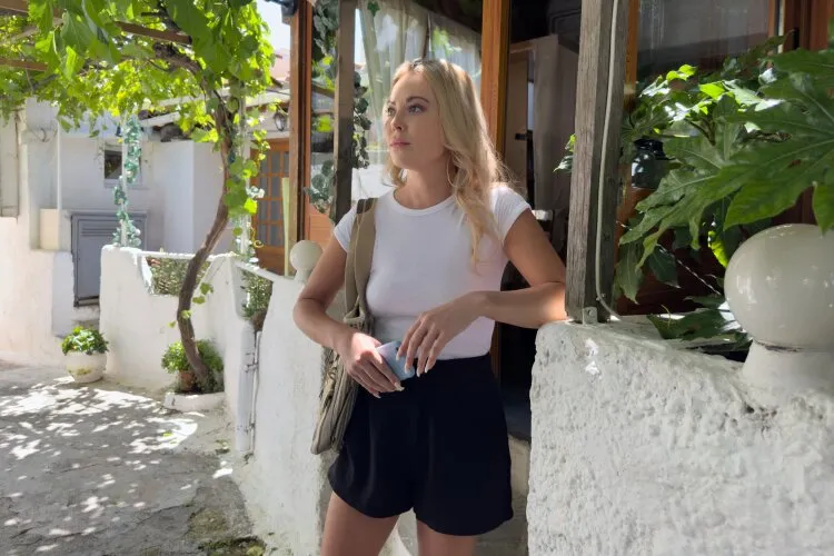 Abby standing thoughtfully in a sunny courtyard, surrounded by lush green plants.