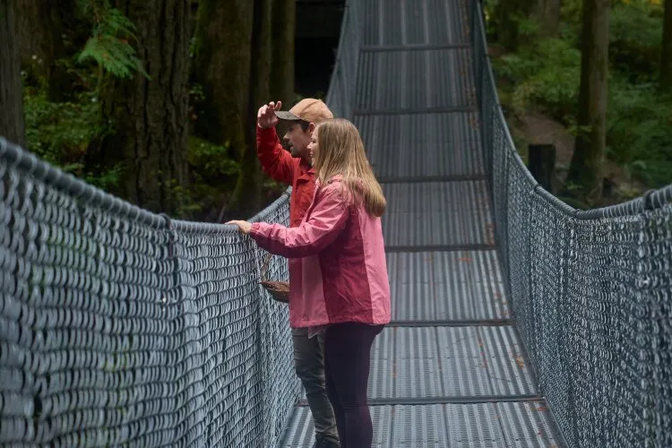 A couple wearing red and pink jackets standing on a suspension bridge, looking out at the scenery in a forest. From Hallmark's "A Whitewater Romance".
