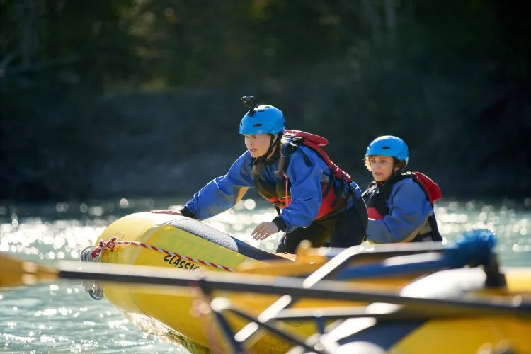 Two people in blue helmets and life jackets maneuvering a yellow raft on a river.