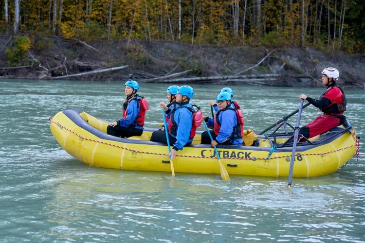  A group of people in blue helmets and life jackets paddling a yellow raft on a river, with a guide at the back steering, surrounded by autumn trees.