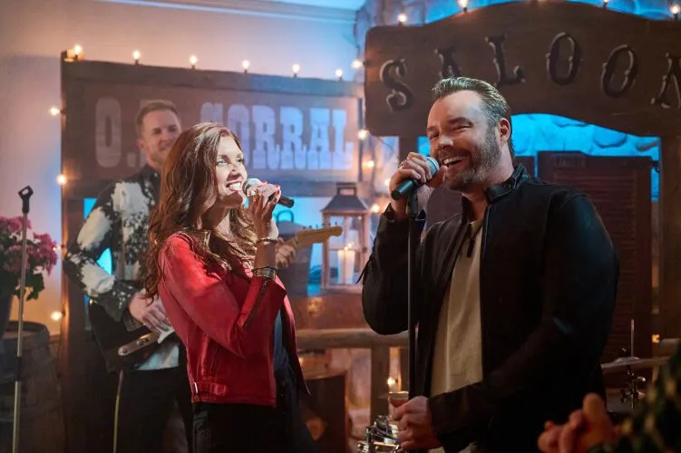A man and a woman singing into microphones at a lively indoor event, with a band playing in the background. From Hallmark's "Big Sky River: The Bridal Path" 