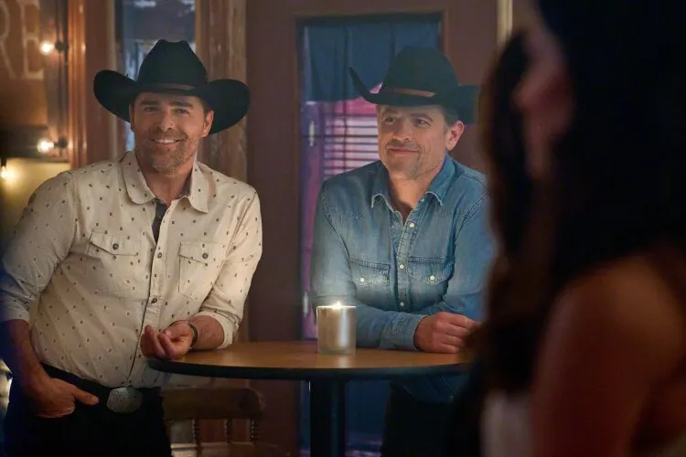 Two men wearing cowboy hats standing inside a dimly lit venue, smiling at someone off-camera.