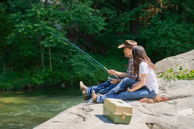A man and a woman sitting on a rock by a river, fishing and enjoying the scenery. From Hallmark's "Big Sky River: The Bridal Path" 