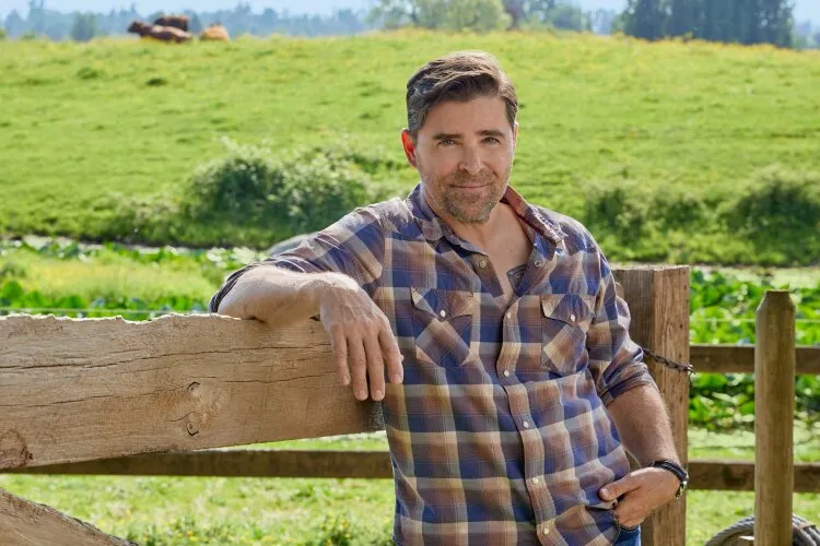 A man in a plaid shirt leaning against a wooden fence, with green fields in the background. From Hallmark's "Big Sky River: The Bridal Path" 