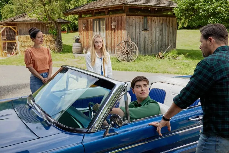 A young man sitting in a convertible car, talking to two women and a man standing outside.