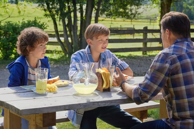A man and two children sitting at an outdoor table, talking and enjoying lemonade. From Hallmark's "Big Sky River: The Bridal Path" 