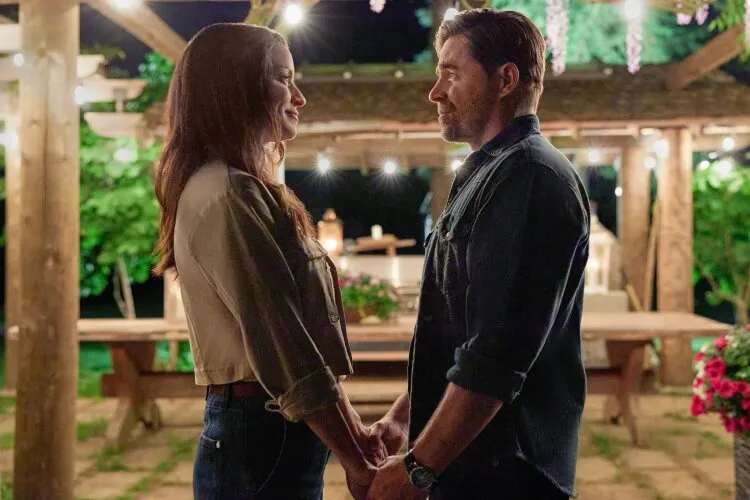 A man and a woman standing outside at night, holding hands and looking at each other lovingly.