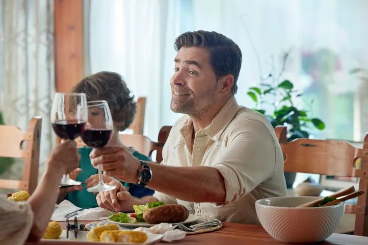 A man sitting at a dinner table, raising a glass of wine with a big smile. From Hallmark's "Big Sky River: The Bridal Path" 