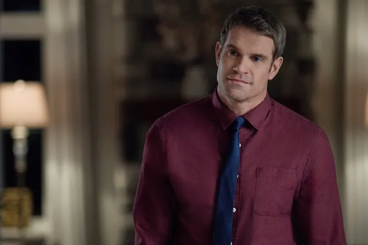 A man in a red shirt and tie standing indoors with a thoughtful expression.