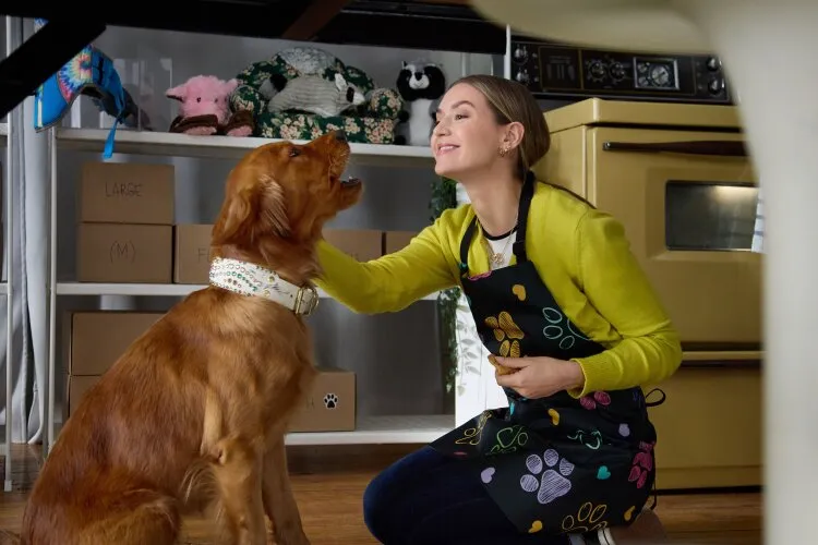 A woman in an apron petting a golden retriever, both looking happy in a kitchen filled with dog supplies.