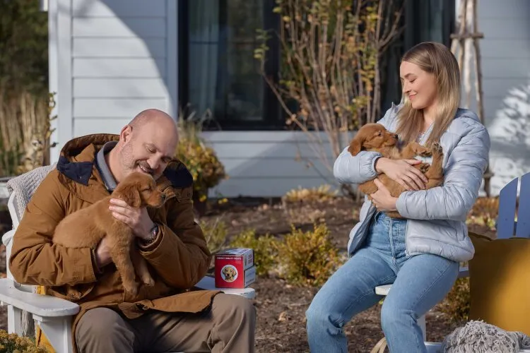 A man and a woman sitting outdoors, each holding a golden retriever puppy and smiling.