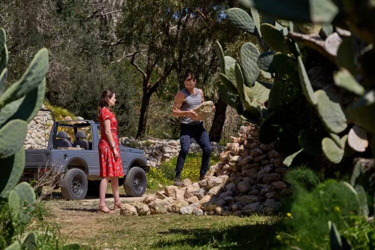  A man lifting a stone while a woman watches, with a jeep and a stone wall in the background.
