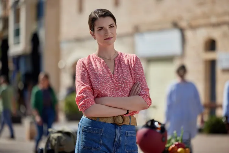  A woman in a red blouse standing with her arms crossed in a town square. From Hallmark's "For Love & Honey"
