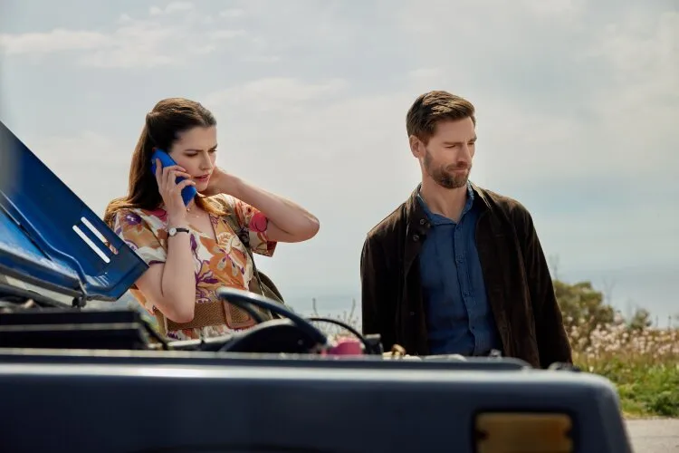  A woman talking on the phone while a man stands beside her, with a broken-down vehicle in the background.