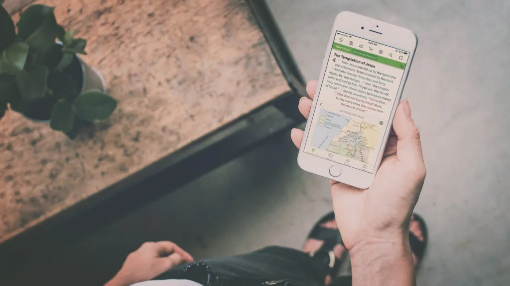 A person holding a smartphone displaying the Olive Tree Bible App, showing a Bible verse with study notes and a map of ancient Israel.