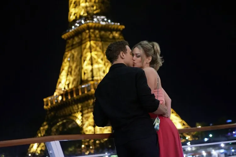 A couple embracing and sharing a kiss on a boat with the illuminated Eiffel Tower in the background at night.