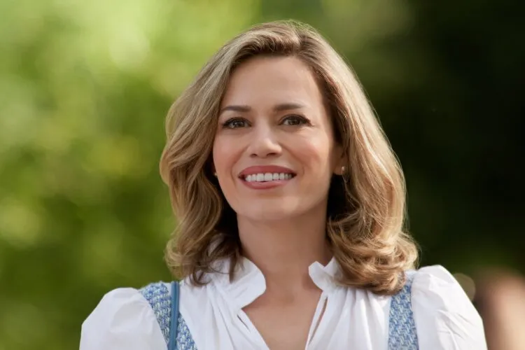 A woman with shoulder-length blonde hair, smiling warmly with greenery in the background.
