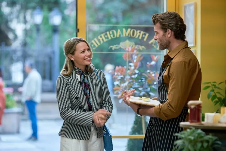 A woman smiling and talking to a man in an apron inside a cheese shop. The shop's sign can be seen through the window, and the interior has a warm, inviting atmosphere.