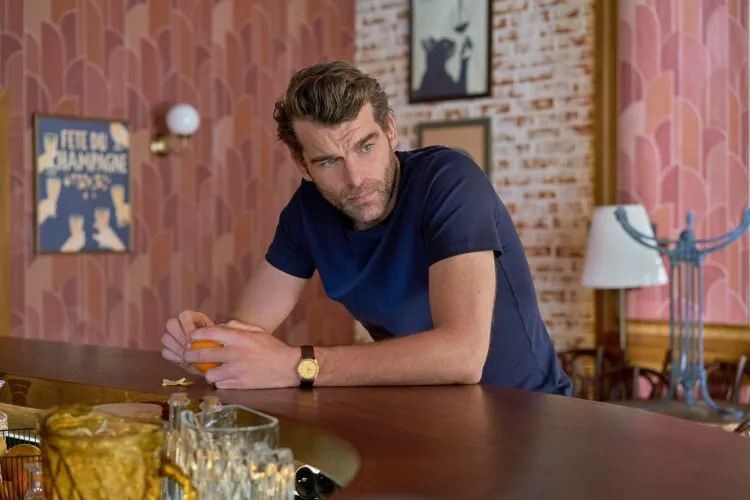A man leaning on a bar counter, looking thoughtful, with a colorful and cozy bar interior in the background.