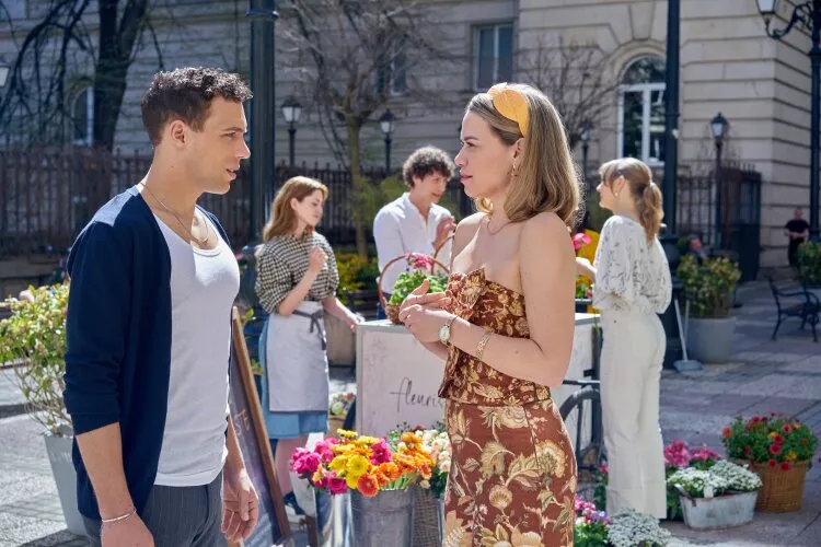 A man and a woman having a conversation in an outdoor market with flower stalls around them. The setting is lively with people and vibrant flowers in the background.