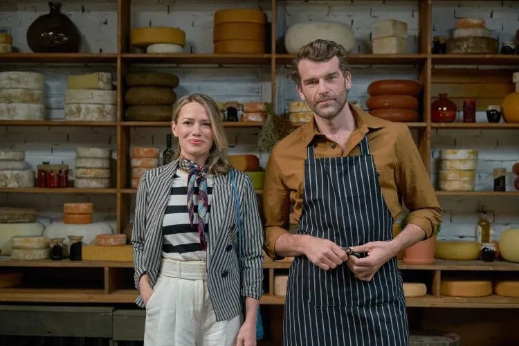 A man in an apron standing next to a woman inside a cheese shop, posing for the camera. The shop's shelves are filled with a variety of cheeses, creating a cozy and rustic atmosphere.