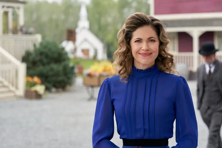 A woman with curly hair, wearing a blue dress, smiling at the camera with a town square in the background.