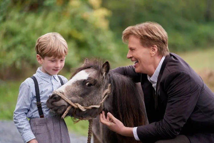 A man in a suit, crouching down and smiling at a young boy with blonde hair who is holding the reins of a small horse. From Hallmark's Truth Be Told When Calls the Heart.