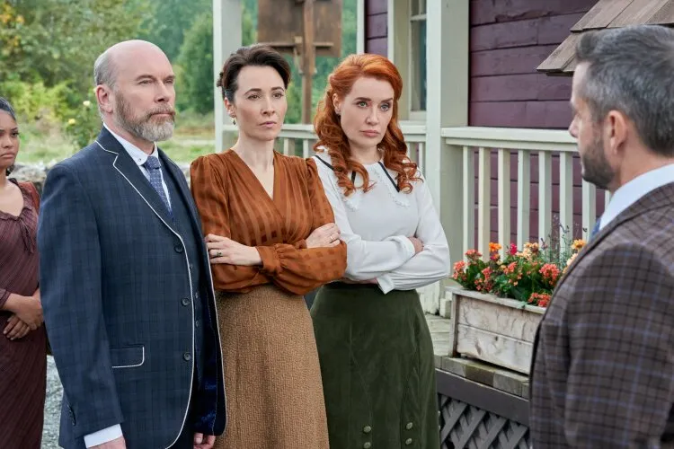 Three people standing with arms crossed, two women and a man, looking serious as they face another man in a suit, in front of a rustic building