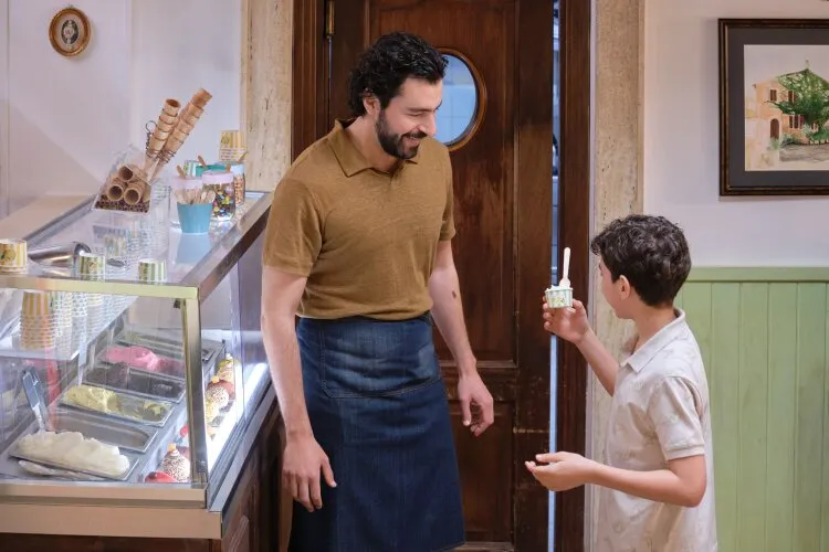 A man behind an ice cream counter smiling at a young boy holding a cup of ice cream.