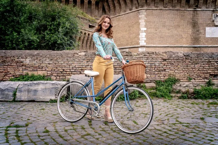A woman in a blue floral top, posing with a bicycle in front of an old brick wall.  "Two Scoops of Italy"  on Hallmark.