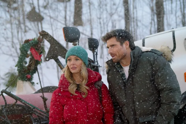 This image shows a man and a woman in a snowy setting, both looking off to the side with expressions of concern or anticipation. The woman is dressed warmly in a red parka with a fur-lined hood and a teal knit beanie, emphasizing the cold weather. The man, similarly dressed in a heavy grey coat with a fur-lined hood, appears engaged in a conversation or observing something out of frame. They are standing in front of a white van decorated with a festive wreath, adding to the wintry, Christmas vibe of the scene. Snowflakes fall gently around them, contributing to the serene yet chilly atmosphere.