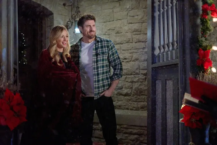 
This image captures a joyful holiday scene featuring a couple standing at the entrance of a beautifully decorated home. The woman, wrapped in a rich red cloak, smiles brightly next to a man dressed casually in a plaid green shirt and jeans. They both exude a relaxed and happy demeanor, clearly enjoying the festive atmosphere. The doorway behind them is adorned with vibrant red poinsettias and greenery, while a light snowfall adds a magical touch to the scene, enhancing the Christmassy feel of the moment.