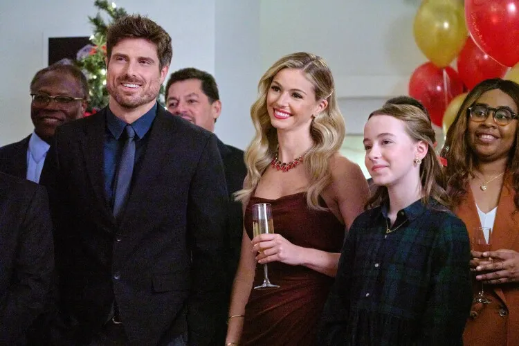 In this image, a group of people are gathered at a festive event, radiating joy and celebration. The focus is on a man and two women, all dressed elegantly for the occasion. The man, in a dark suit, stands with a relaxed, pleased expression, accompanied by a woman in a stunning burgundy dress with a radiant smile. To their side, a younger girl in a dark plaid dress shares in the joyful moment. The background, decorated with Christmas trees and balloons, suggests a holiday party or a similar celebratory gathering. Their expressions and the warm atmosphere convey a sense of happiness and togetherness typical of festive celebrations.