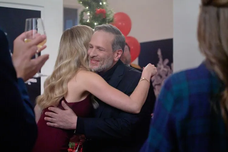 This image depicts a heartfelt moment at a festive celebration. An older man, dressed in a sleek black suit, embraces a younger woman wearing an elegant burgundy dress. His expression is one of genuine affection and joy as he smiles warmly during the embrace. The background is lively with other guests and holiday decorations, including balloons and a Christmas tree, enhancing the festive atmosphere of the scene. This intimate gesture, set against the backdrop of a holiday party, captures the spirit of familial love or close friendship during the celebratory season.