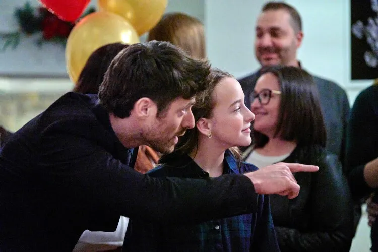
This image captures a lively and engaging moment at a party. A man in a dark blazer playfully points towards something out of frame, drawing the attention of a young girl beside him. She looks on with a smile, intrigued by whatever he is showing her. Both are surrounded by other guests, contributing to the cheerful and sociable atmosphere of the event. The man's dynamic gesture and the girl's attentive expression suggest a fun and interactive environment, likely filled with storytelling or sharing amusing observations.