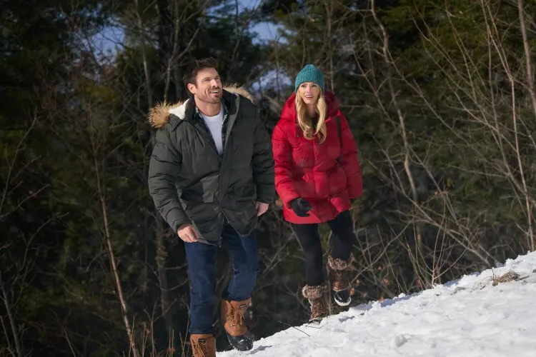 In this image, a man and a woman are depicted enjoying a sunny winter day outdoors. The couple, both dressed warmly in winter attire, are walking and laughing together on a snow-covered hill. The man wears a green parka and jeans, while the woman is in a vibrant red jacket and a teal beanie, which adds a cheerful pop of color against the snowy landscape. Their expressions of joy and the active setting suggest a playful, carefree moment shared between them, capturing the essence of a pleasant winter outing.