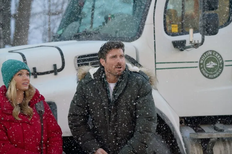 This image features a man and a woman standing in a snowy setting, both looking upwards with expressions of awe or interest. The man, dressed in a grey parka with a fur-lined hood, seems to be reacting to something out of frame, while the woman, in a red parka and teal beanie, also gazes in the same direction with a look of pleasant surprise. They are standing next to a white truck adorned with a green and white logo, indicating they might be part of some outdoor activity or event. The falling snow adds to the winter ambiance, creating a scene that feels both adventurous and festive.