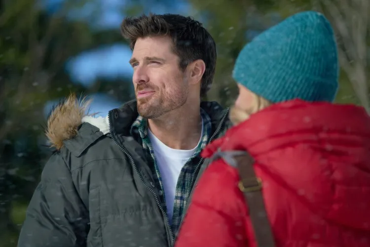 This image captures a man in a contemplative moment outdoors during winter. He is dressed in a gray winter jacket with a fur-lined hood and a plaid shirt, suggesting a casual but cold setting. Beside him, part of a woman in a red jacket can be seen, indicating they are together. The man is looking off into the distance with a thoughtful expression, perhaps observing the natural winter landscape or engaged in a deep conversation. The scene is set against a backdrop of snowflakes gently falling, adding to the serene and reflective mood of the moment.