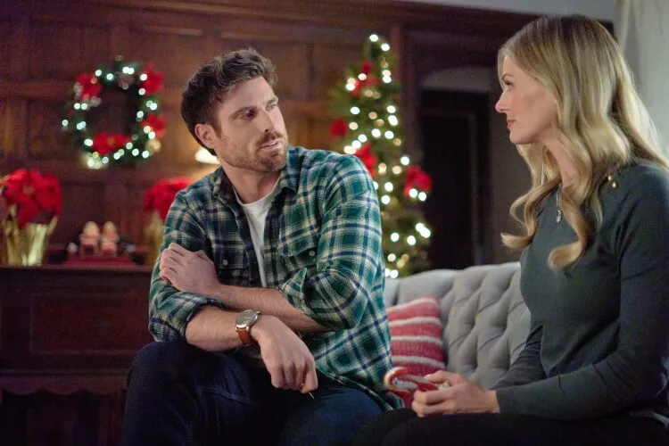 This image captures a heartfelt indoor conversation between a man and a woman, set against a cozy holiday backdrop. The man, dressed casually in a plaid shirt, leans forward with a thoughtful expression, engaging intently with the woman. She, dressed in a simple grey top, listens carefully, indicating a serious or meaningful dialogue. Behind them, the room is warmly decorated with Christmas lights and poinsettias, enhancing the intimate and festive atmosphere. This scene likely portrays a moment of personal connection or discussion, set during the holiday season.