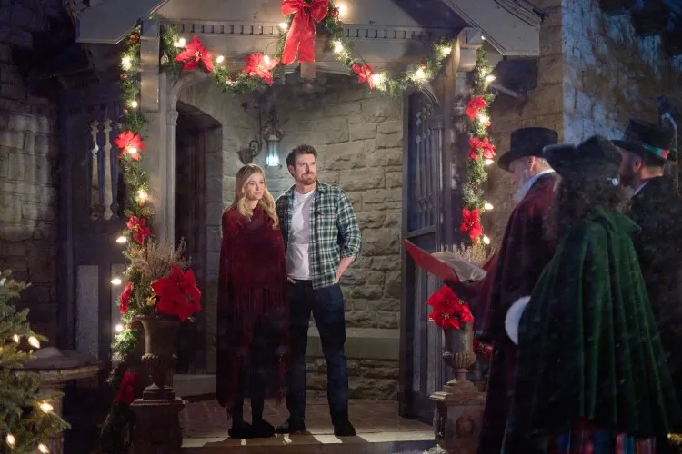 In this image, a couple stands at the entrance of a stone house adorned with festive Christmas decorations, including red poinsettias and wreaths. The woman, wrapped in a dark red cloak, and the man, in casual attire with a plaid shirt, both look outwards with welcoming expressions, perhaps greeting guests. The setting is enchanting with its twinkling lights and traditional holiday decor, creating a warm and inviting atmosphere. This scene perfectly captures the spirit of the season, portraying a moment of shared joy and anticipation during a festive event.