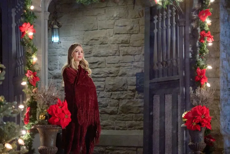 This image features a woman standing alone on a beautifully decorated porch adorned with festive Christmas decorations. She is wrapped in a luxurious dark red cloak, looking thoughtfully into the distance. The setting is warmly lit, with twinkling lights and vibrant red poinsettias enhancing the festive atmosphere. The stone walls of the porch and the traditional wooden door further add to the old-world charm of the scene. Her reflective pose and the serene setting evoke a sense of quiet contemplation amidst the festive surroundings, suggesting a moment of personal reflection during the holiday celebrations.






