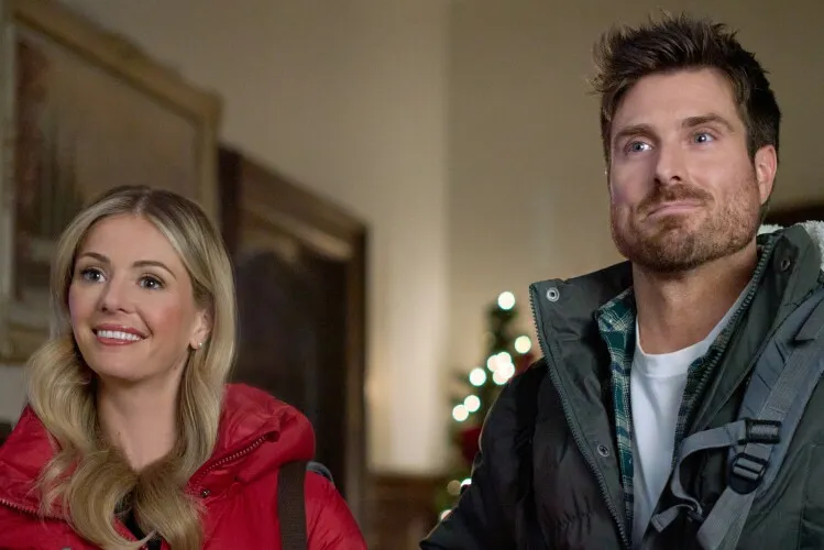 This image captures a moment of joyful anticipation between a man and a woman, likely during a holiday event. The woman, dressed in a bright red jacket, smiles broadly, her face lit with happiness. Beside her, the man, wearing a practical winter jacket, looks upwards with an expression of wonder and curiosity, as if responding to something intriguing or delightful out of view. Their expressions and the cozy indoor setting, adorned with Christmas lights and holiday decor, suggest a warm, festive atmosphere filled with cheerful interactions.