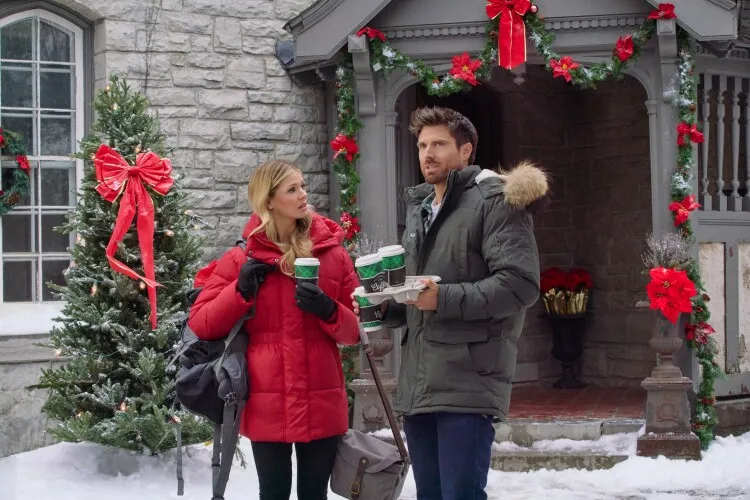 In this image, a couple stands outside a beautifully decorated home adorned with Christmas wreaths and red bows. The man, dressed warmly in a green parka and carrying a tray with coffee cups, seems to be in a conversation with the woman, who is dressed in a red winter jacket. She looks slightly puzzled or curious, perhaps in response to something the man is explaining or pointing out. The snowy setting and festive decorations create a quintessential holiday scene, capturing a moment of casual interaction in the midst of a winter day.