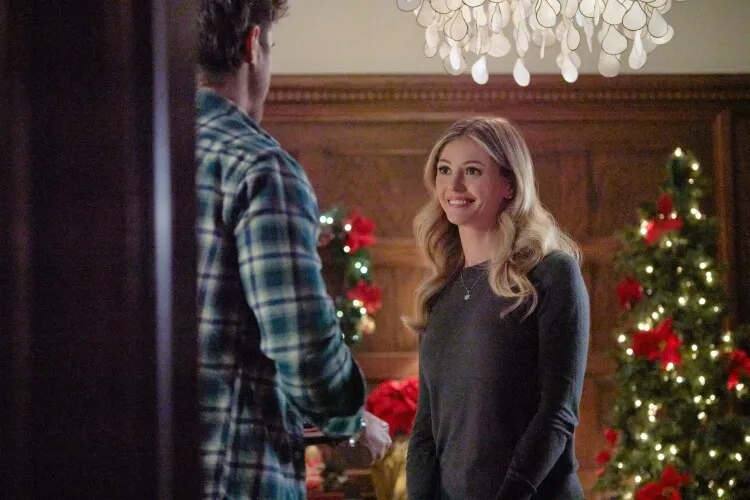
In this image, we see a woman, brightly smiling and facing a man whose back is partially shown to the camera. The setting is a beautifully decorated room, suggestive of a festive season with its richly adorned Christmas tree and elegant chandelier overhead. The woman, dressed casually in a grey top, looks delighted, possibly in the midst of a friendly or intimate conversation. The warmth and joy evident in her expression reflect a cozy, holiday atmosphere, perfect for heartfelt exchanges and festive gatherings.