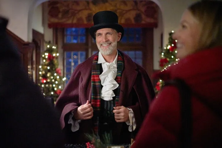 This image captures a charming holiday scene featuring an older man dressed in festive attire. He's wearing a dark burgundy cape over a white shirt with a ruffled front, a plaid scarf, and a classic black top hat, creating a Dickensian or Victorian-era holiday look. His warm smile and the joyful twinkle in his eyes suggest he's playing a welcoming host or a character in a holiday event. The background, adorned with a beautifully decorated Christmas tree and lit-up windows, enhances the festive mood of the setting, making it feel like a scene from a classic holiday story.