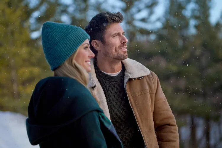 
In this image, a man and a woman are captured in a serene winter moment, surrounded by gently falling snowflakes. The woman, wearing a teal beanie and a green scarf, smiles as she looks into the distance, her profile hinting at a joyful demeanor. The man, dressed in a brown leather jacket with a shearling collar over a dark knit sweater, gazes off into the distance with a contemplative expression. This scene conveys a sense of companionship and warmth amid the chilly, picturesque landscape, suggesting a moment of shared appreciation for the winter scenery around them.