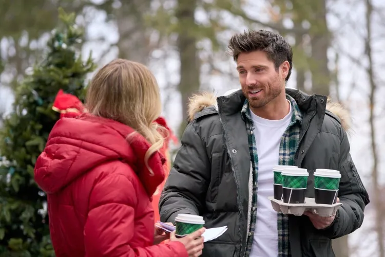 
This image captures a casual winter moment between a man and a woman. The man, dressed warmly in a green plaid shirt and heavy grey parka with a fur-lined hood, holds a tray with three cups of coffee, offering one to the woman. He smiles warmly at her, suggesting a friendly or intimate exchange. The woman, facing away from the camera, is clad in a vibrant red parka, adding a pop of color to the snowy scene. This snapshot exudes a cozy, cheerful vibe, emphasizing a simple, enjoyable interaction on a cold day.