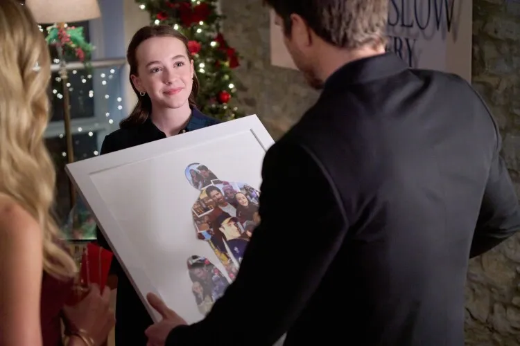 In this image, a young woman, likely at a holiday party, graciously accepts a large framed photograph as a gift. She is dressed in a simple, elegant black outfit and her expression is one of happiness and gratitude as she looks towards the man presenting the gift, whose back is to the camera. The framed photograph features a collage of people, suggesting it holds sentimental value, likely capturing moments with friends or family. The setting, adorned with Christmas decorations, adds to the warm and festive atmosphere of the occasion.