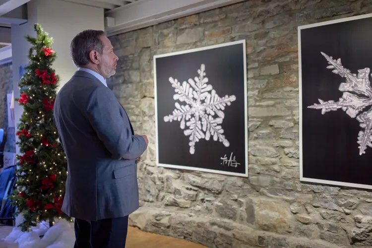 In this image, a distinguished middle-aged man in a suit is thoughtfully admiring two large framed artworks featuring intricate snowflake designs on a stone wall. The artwork on the left showcases a detailed white snowflake on a black background, while the piece on the right presents a more abstract and dynamic snowflake design. Positioned next to a decorated Christmas tree, the setting evokes a festive atmosphere, blending traditional holiday decor with contemporary art. The man's contemplative stance suggests he is either an art enthusiast or someone deeply appreciating the festive spirit conveyed through the artwork.